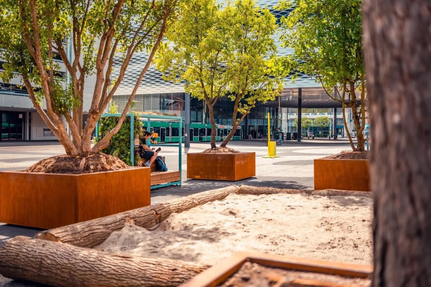 Eine öffentliche Freifläche mit Bäumen in großen quadratischen Pflanzgefäßen, einer von Baumstämmen gesäumten Sandfläche, einer Person, die auf einer Bank sitzt, und einem modernen Glasgebäude im Hintergrund.