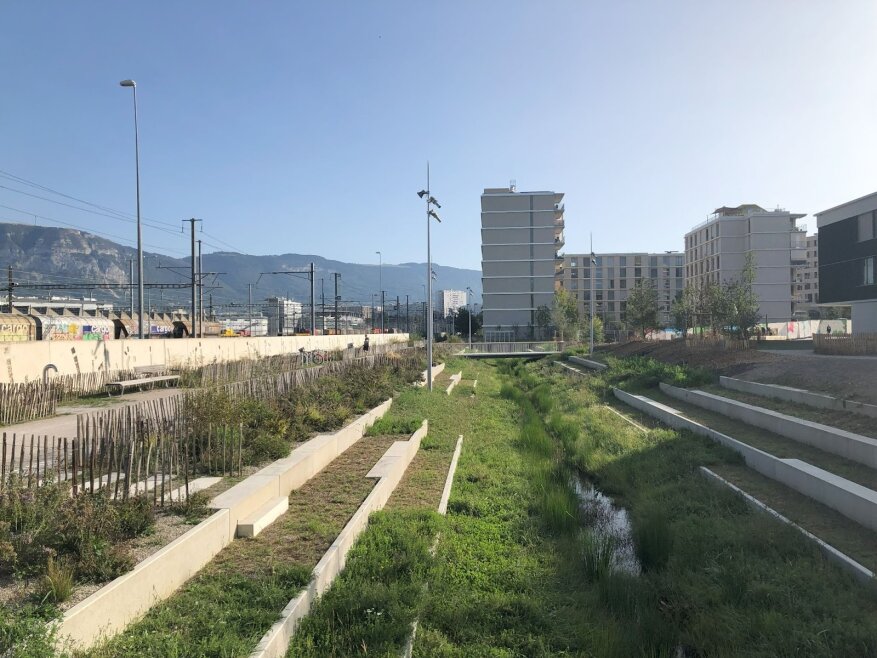 Ein moderner Stadtpark mit abgestuften Sitzplätzen, einem schmalen Bach und verstreuten kleinen Bäumen. Im Hintergrund sind Wohnhäuser und Bahngleise zu sehen und die Berge unter einem klaren Himmel.