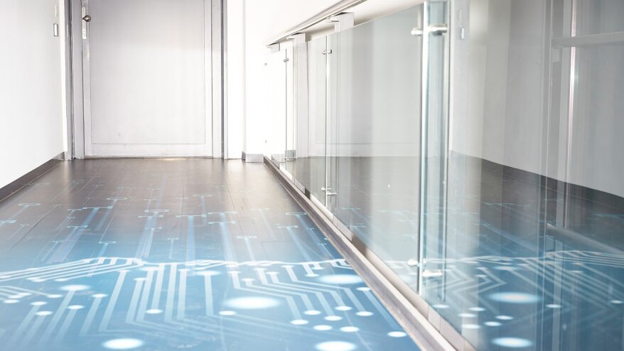 Ein moderner Flur mit einem Glasgeländer und einem grauen Boden mit blauem Leiterplattenmuster, der zu einer geschlossenen hellgrauen Tür führt. Der Bereich ist durch natürliches Licht hell erleuchtet.