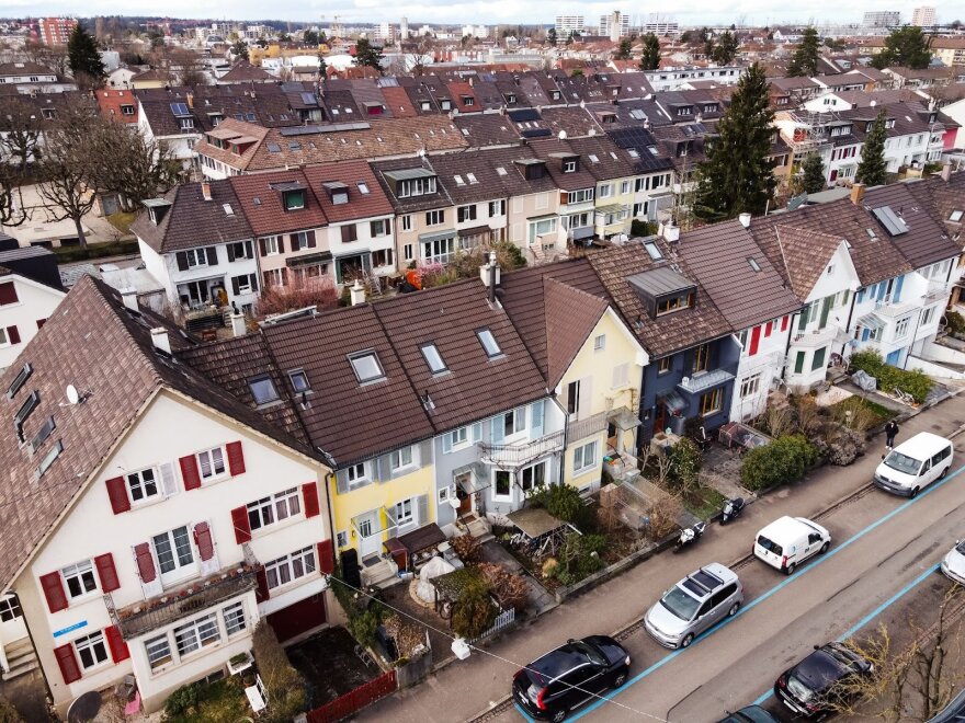 Luftaufnahme einer Wohnstraße mit bunten, angebauten Häusern mit schrägen Dächern, parkenden Autos entlang der Straße und Reihen ähnlicher Häuser, die sich in die Ferne erstrecken.