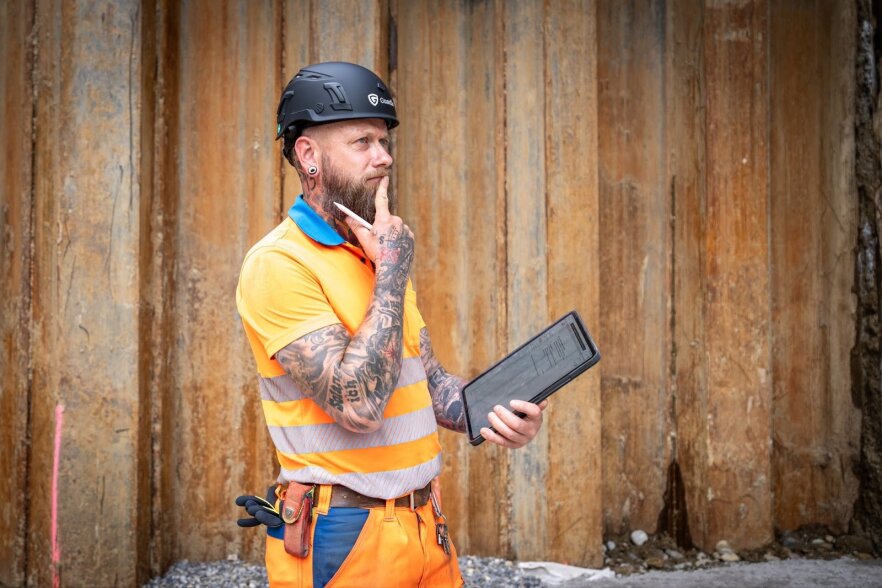 Ein tätowierter Bauarbeiter in einer orangefarbenen Sicherheitsweste und einem Schutzhelm hält eine Tafel in der Hand und steht nachdenklich mit der Hand am Kinn vor einer rostigen Metallwand.