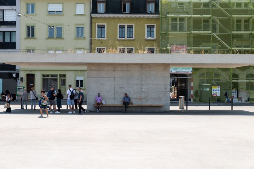 An einem sonnigen Tag wartet eine Gruppe von Menschen an einer modernen Bushaltestelle aus Beton mit einem grün überdachten Wartehäuschen vor pastellfarbenen Wohnhäusern. Einige sitzen auf der Bank, andere stehen, und die Straße ist ruhig.