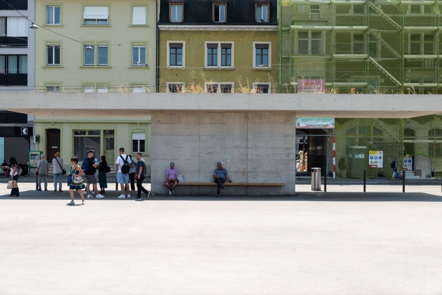 An einem sonnigen Tag wartet eine Gruppe von Menschen an einer modernen Bushaltestelle aus Beton mit einem grün überdachten Wartehäuschen vor pastellfarbenen Wohnhäusern. Einige sitzen auf der Bank, andere stehen, und die Straße ist ruhig.