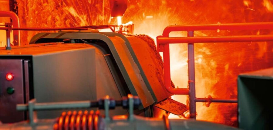 In einer hellen, feurigen Szene in einem Stahl- oder Metallverarbeitungswerk ergießt sich geschmolzenes Metall aus Industriemaschinen, wobei Funken und intensives orangefarbenes Licht die Anlagen beleuchten.