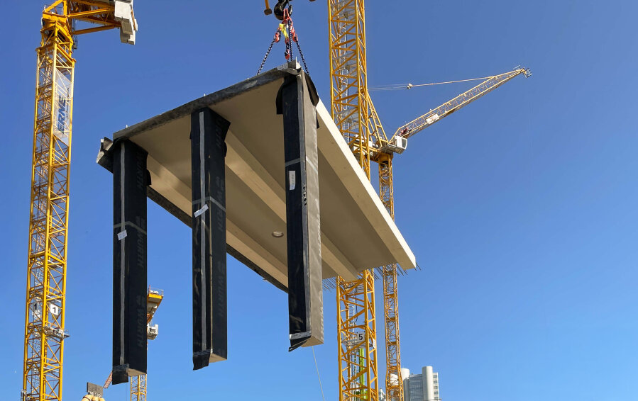 Eine große Betonstruktur mit drei schwarzen Stützen wird von einem gelben Baukran vor einem klaren blauen Himmel angehoben. Im Hintergrund sind zwei weitere Kräne und ein hohes Gebäude zu sehen.