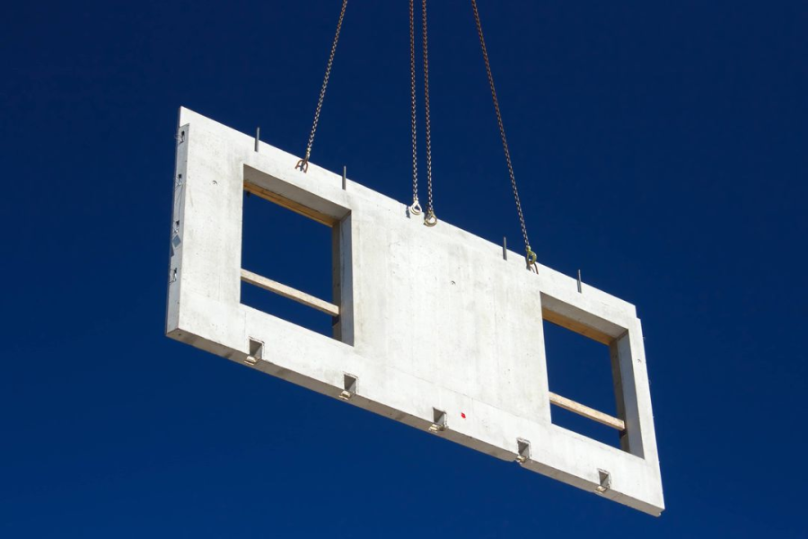 Eine große Betonwandtafel mit zwei rechteckigen Fensteröffnungen hängt an Seilen in der Luft vor einem klaren blauen Himmel.