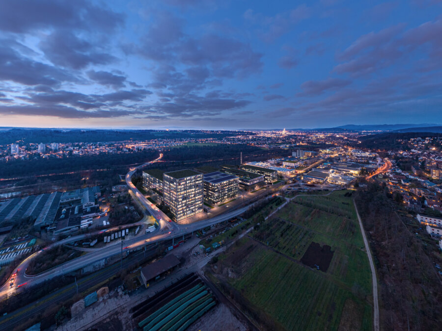 Luftaufnahme eines modernen Bürokomplexes mit beleuchteten Gebäuden in der Abenddämmerung, umgeben von Feldern, Straßen und einer sich in die Ferne erstreckenden Stadtlandschaft unter einem teilweise bewölkten Himmel.