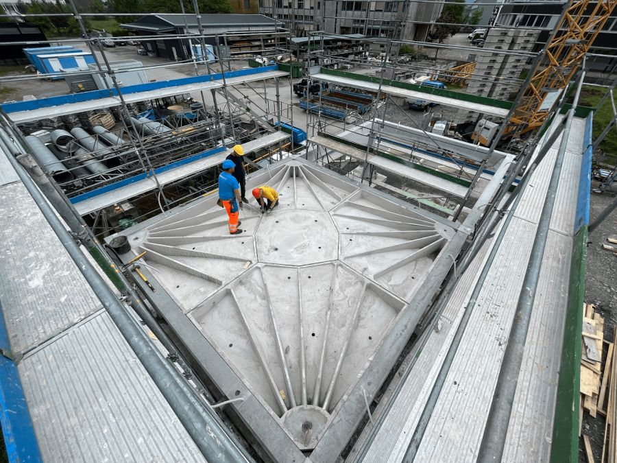 Zwei Bauarbeiter in Schutzkleidung stehen auf einer großen Betonstruktur mit radialen Stützen auf einer Baustelle, umgeben von Gerüsten, Geräten und Industriematerialien.