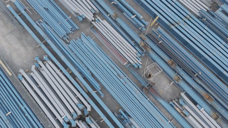 Luftaufnahme von ordentlich angeordneten Stapeln langer blauer Stahlträger und -rohre auf einer Baustelle, mit sichtbarem Betonboden zwischen den geordneten Reihen.