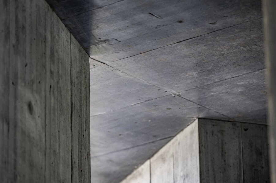 Eine Nahaufnahme von sich kreuzenden Betonwänden und -decken, die geometrische Linien, Winkel und Texturen in einer modernen architektonischen Struktur zeigen. Schatten heben die glatten, grauen Oberflächen hervor.