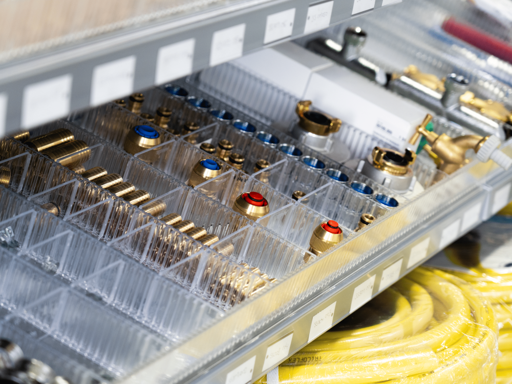 Eine Nahaufnahme von organisierten Messingarmaturen mit roten und blauen Markierungen in durchsichtigen Plastikfächern in einer Metallschublade, darunter sind aufgerollte gelbe Schläuche zu sehen.