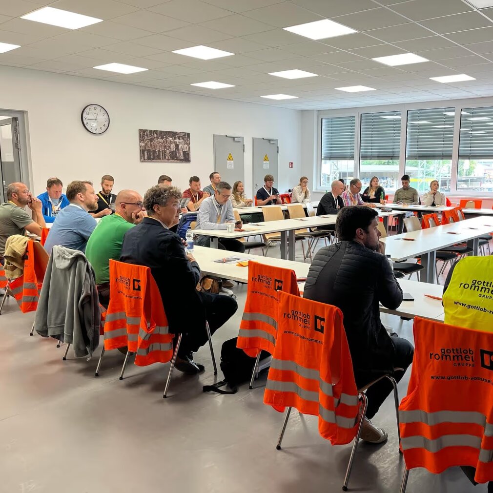 Die Menschen sitzen in einem Besprechungsraum, einige tragen orangefarbene Sicherheitswesten oder haben diese über die Stühle gezogen. Die Tische sind U-förmig angeordnet, und an der Wand hängen eine Uhr und ein Foto. Große Fenster lassen natürliches Licht herein.