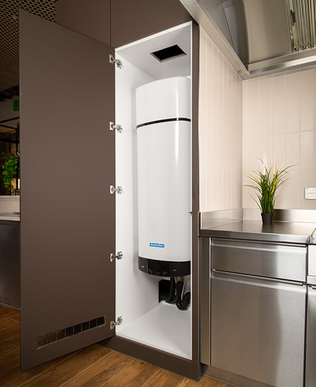 Ein weißer Warmwasserbereiter ist in einem hohen, offenen Schrank in einer modernen Küche mit braunen und Edelstahl-Oberflächen installiert. Eine kleine Grünpflanze steht auf der Arbeitsplatte neben dem Schrank.