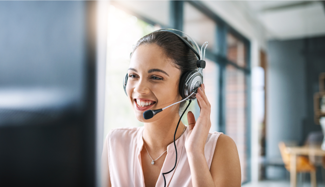 Eine lächelnde Frau mit Headset sitzt an einem Schreibtisch in einem hellen Büro und blickt beim Sprechen auf einen Computerbildschirm, was darauf schließen lässt, dass sie im Kundendienst oder in einem Callcenter arbeitet.
