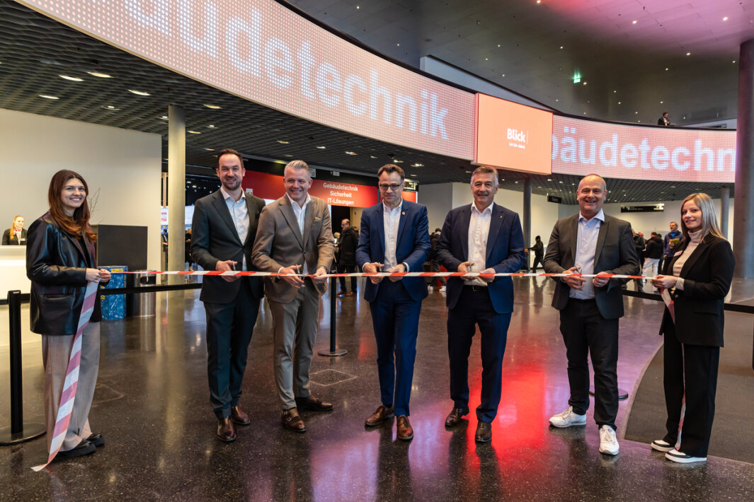 Sieben Personen in Geschäftskleidung stehen in einer Reihe und halten ein feierliches Band in der Hand. Sie lächeln für ein Foto in einem überdachten Veranstaltungsraum mit digitalen Zeichen und einem modernen Design im Hintergrund.