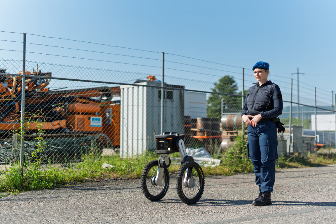 Ein Polizeibeamter in Uniform steht im Freien auf einem gepflasterten Weg neben einem zweir&auml;drigen Roboter. Im Hintergrund sind ein Maschendrahtzaun und Industrieanlagen bei strahlend blauem Himmel zu sehen.