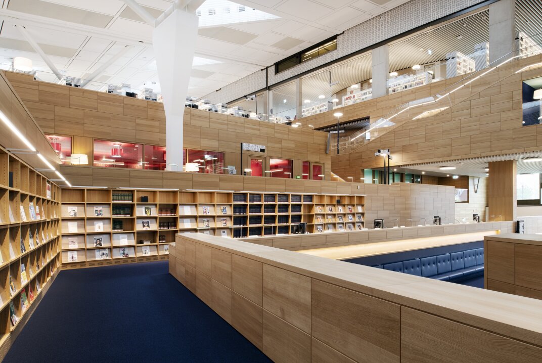 Moderne Bibliothekseinrichtung mit Bücherregalen aus Holz, blauem Teppich und großzügigen Sitzgelegenheiten. Große Fenster und offene Balkone in den oberen Etagen sorgen für natürliches Licht und den Blick auf weitere Bücherregale und Arbeitsbereiche.
