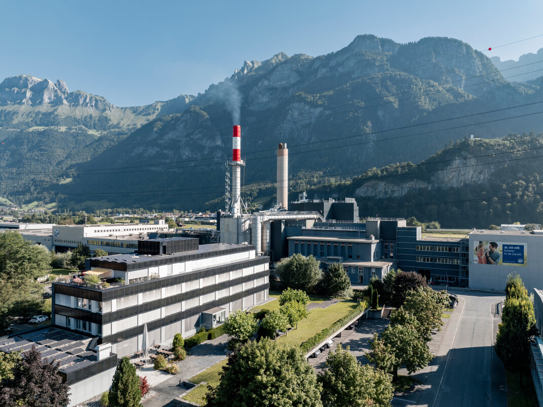 Eine große Industrieanlage mit hohen Schornsteinen liegt vor der Kulisse grüner Berge und eines klaren Himmels, umgeben von Bäumen und modernen Gebäuden an einem sonnigen Tag.