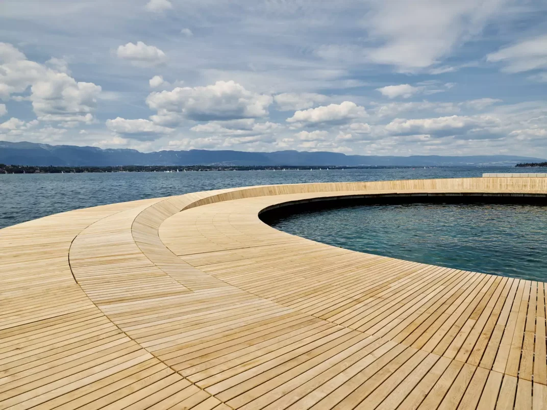 Ein runder Holzsteg, der ein rundes Wasserbecken umgibt, liegt neben einem gro&szlig;en See unter einem teilweise bew&ouml;lkten Himmel mit fernen Bergen im Hintergrund.