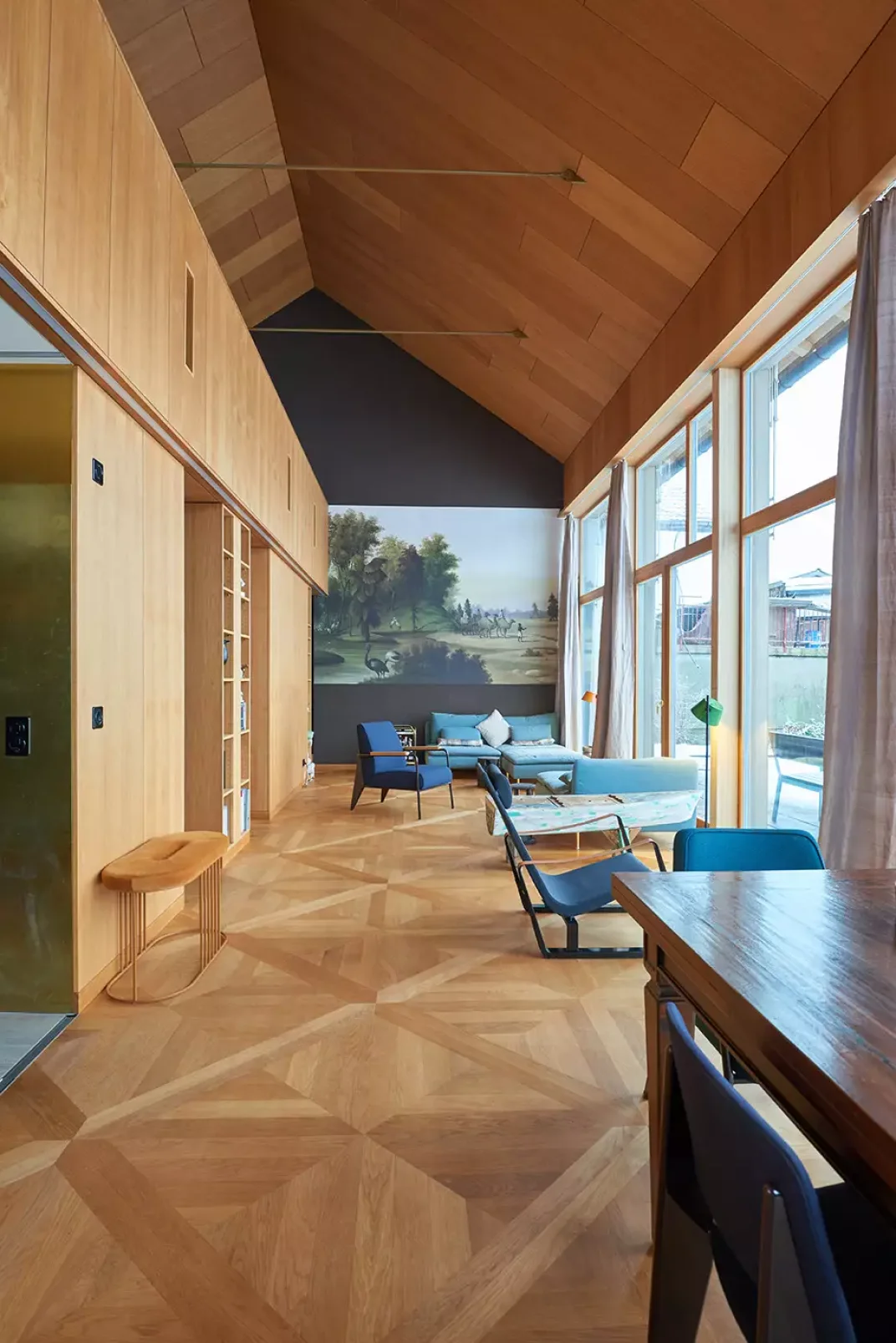 Ein moderner Wohnraum mit hohen Holzdecken, geometrischen Holzb&ouml;den, gro&szlig;en Fenstern mit transparenten Vorh&auml;ngen und modernen M&ouml;beln. Ein gro&szlig;es Wandgem&auml;lde mit einer Parkszene ziert die gegen&uuml;berliegende Wand.