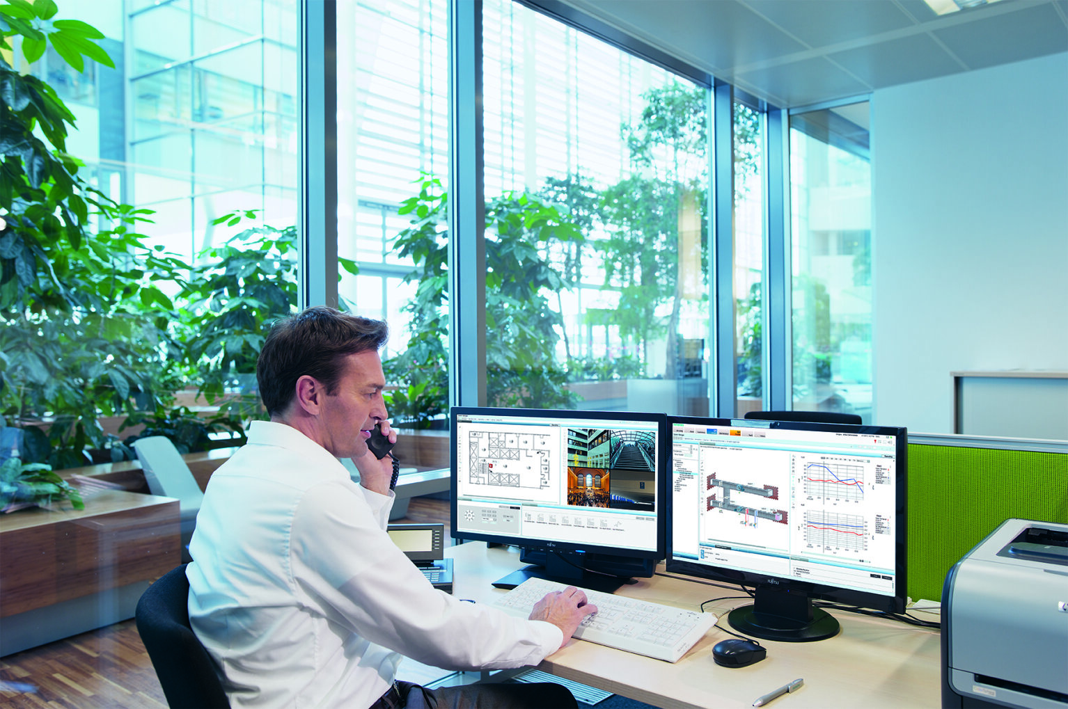 Ein Mann im weißen Hemd sitzt an einem Schreibtisch in einem modernen Büro mit großen Fenstern und vielen Pflanzen. Er telefoniert und arbeitet an einem Computer mit drei Monitoren, auf denen technische Diagramme und Daten angezeigt werden.