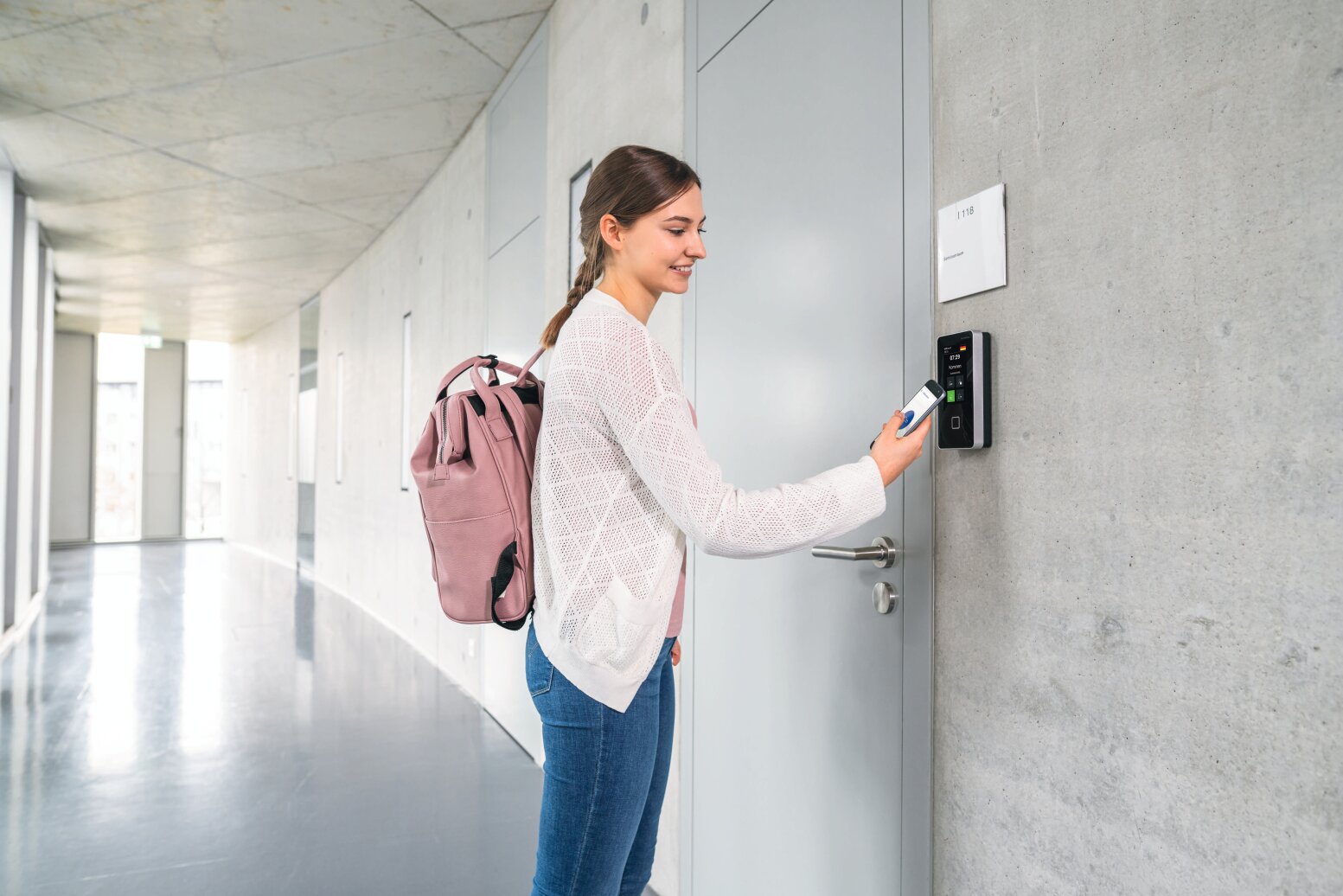 Eine junge Frau mit einem rosa Rucksack öffnet mit einer Zugangskarte eine gesicherte Tür in einem modernen Flur mit grauen Böden und Wänden.