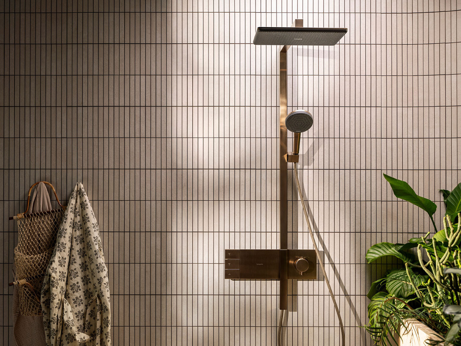Une douche moderne avec une douche à effet pluie carrée et une douchette à main est montée sur un mur carrelé, à côté duquel se trouvent une serviette, un sac tressé et une plante verte en pot. La lumière du soleil éclaire doucement la scène.