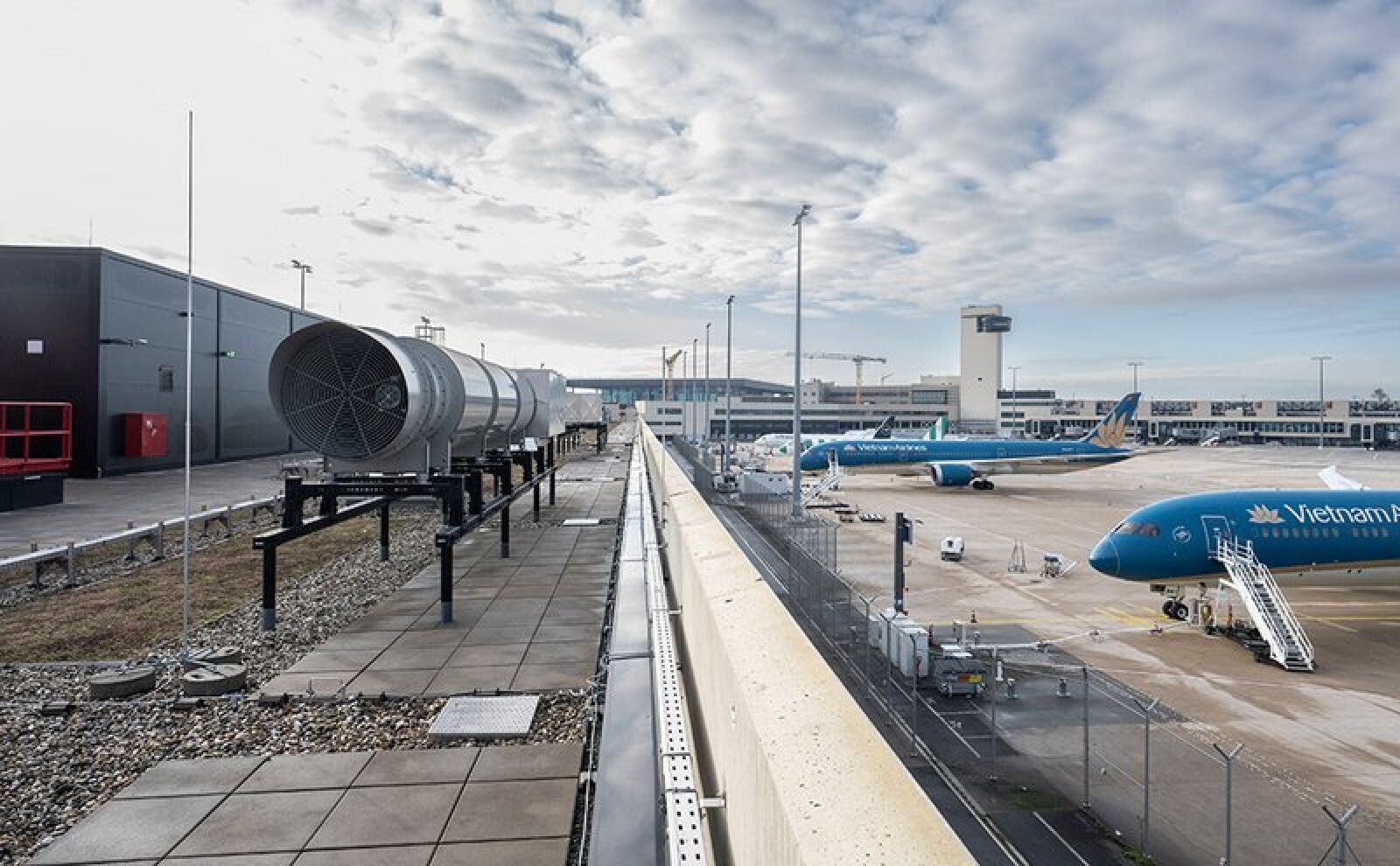 Un tarmac d'aéroport avec des avions de Vietnam Airlines garés aux portes d'embarquement, des chariots à bagages à proximité et un grand système de ventilation sur un toit sous un ciel partiellement nuageux.