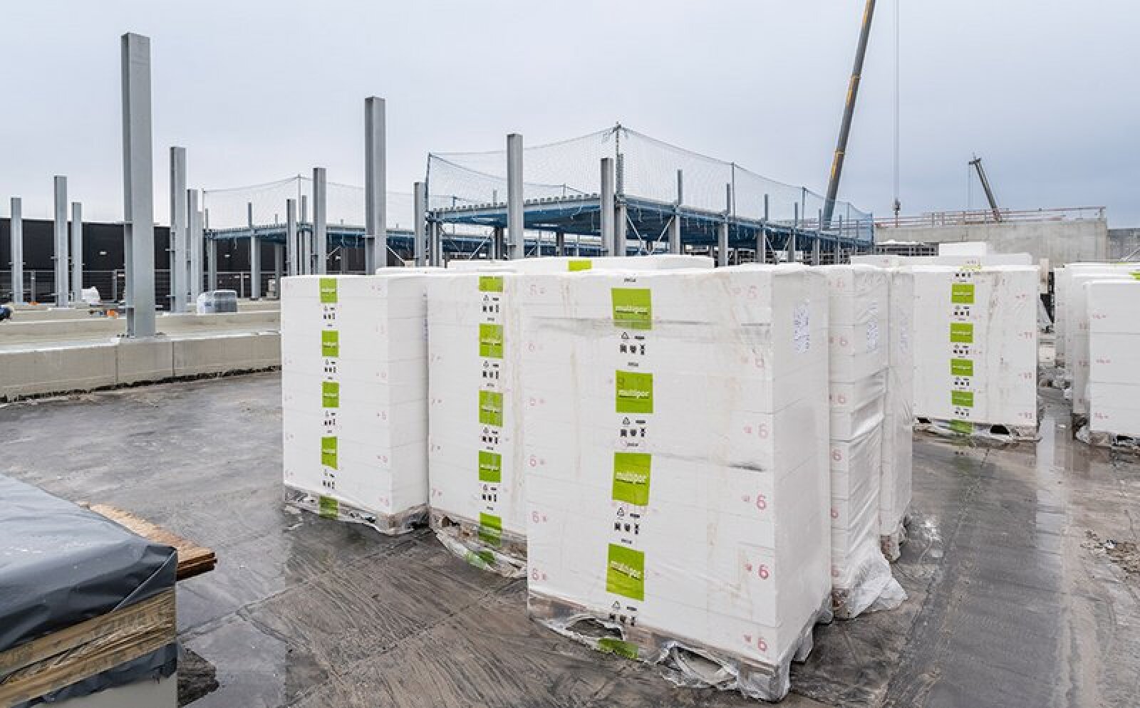 Des piles de panneaux isolants blancs enveloppés de plastique sont posées sur un chantier avec des poutres métalliques, des structures inachevées et une grue en arrière-plan, sous un ciel nuageux.