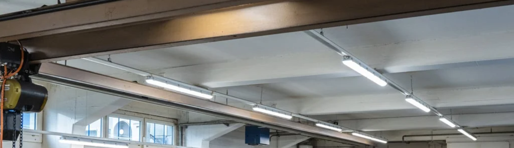 Decke eines Industriegebäudes mit freiliegenden Balken, Leuchtstoffröhren und einigen sichtbaren Rohren und Fenstern an den oberen Wänden.