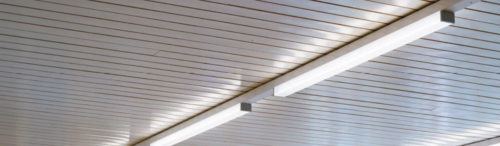 Eine Nahaufnahme einer modernen Decke mit parallelen weißen Paneelen und zwei langen, rechteckigen LED-Leuchten, die quer darüber verlaufen.