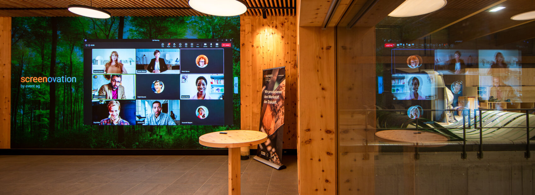 In einem modernen Besprechungsraum mit Holzwänden befindet sich ein großer Bildschirm, auf dem eine virtuelle Videokonferenz mit mehreren Teilnehmern läuft. Von der Decke hängen kreisförmige Leuchten, und im Raum sind runde Holztische aufgestellt.