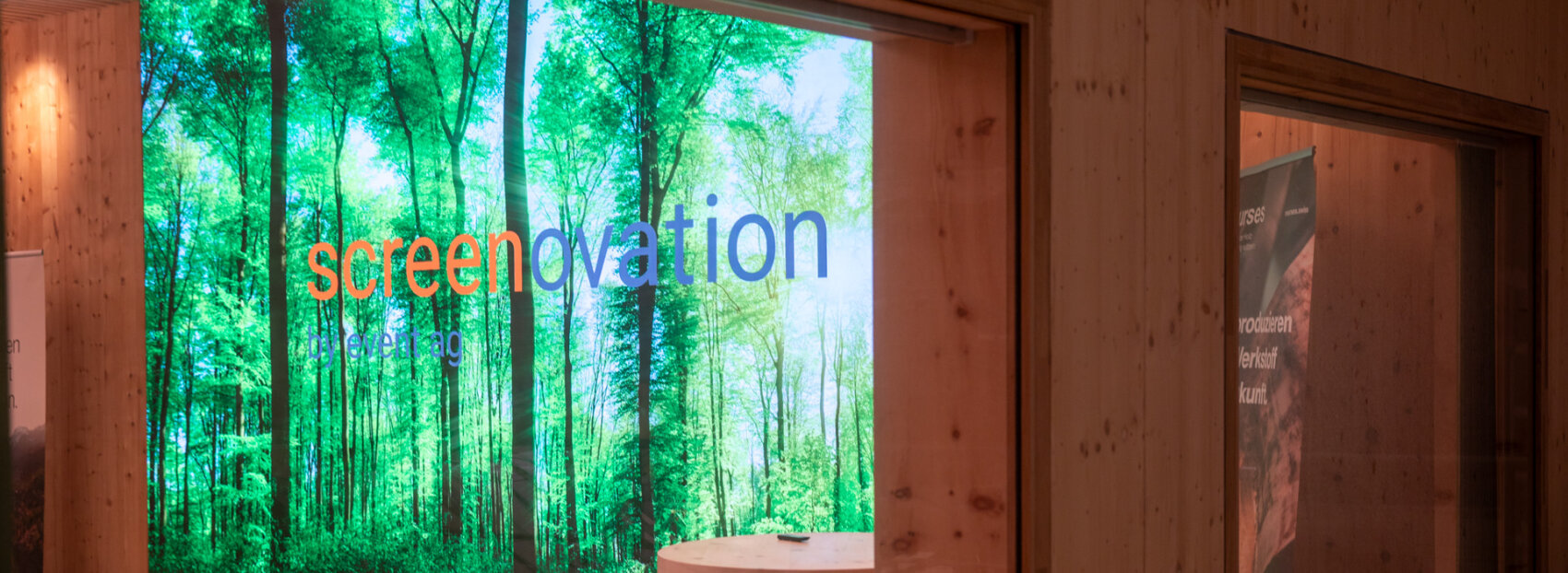 Ein großer digitaler Bildschirm zeigt ein Bild eines grünen Waldes mit dem Text screenovation vor einer Holzwand und einem Fenster in einem modernen Gebäude.