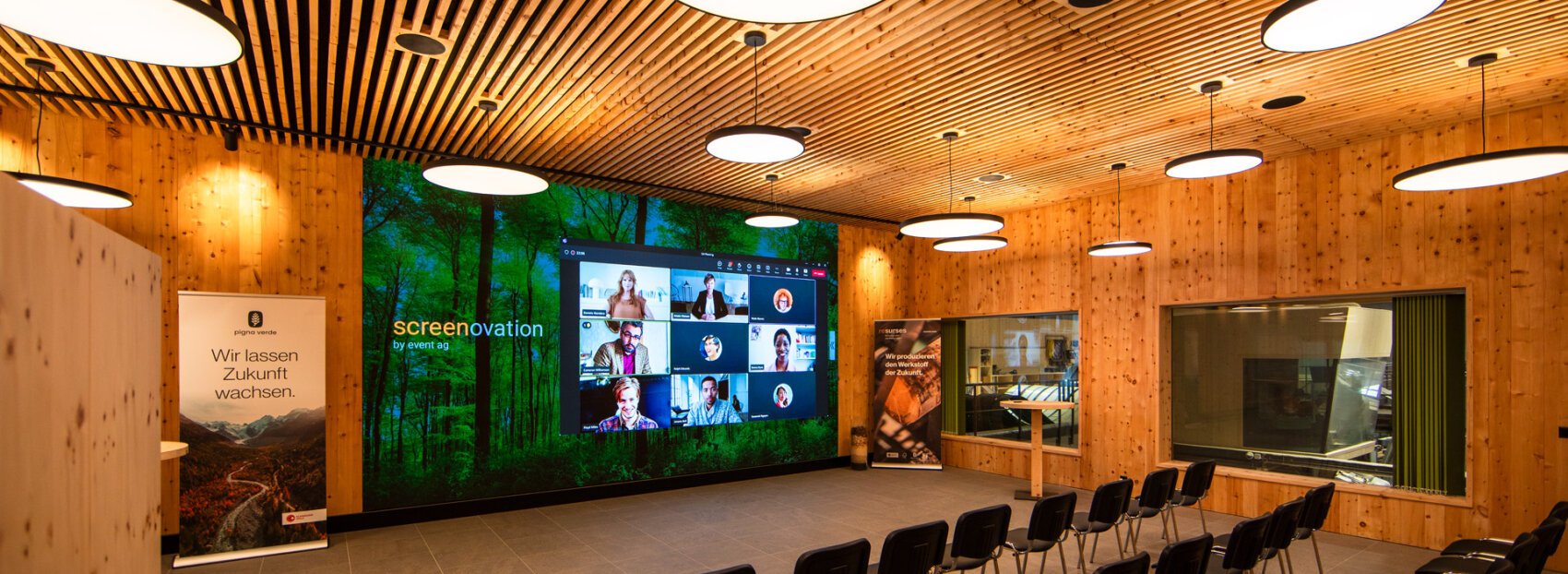 Ein moderner Konferenzraum mit hölzernen Wänden und Decken, kreisförmigen Leuchten, leeren Stühlen, einem großen Bildschirm, auf dem ein Videoanruf angezeigt wird, und Bannern mit deutschem Text.