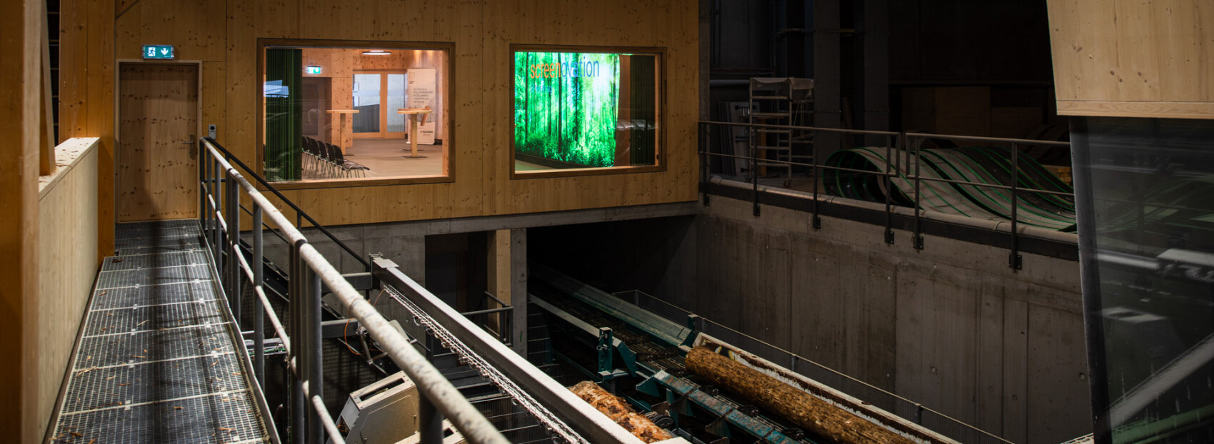 Eine moderne Holzverarbeitungsanlage mit Metallstegen, Holzwänden, großen Fenstern, die den Blick auf einen Raum im Inneren freigeben, und Stämmen, die auf einem Förderband nach unten transportiert werden.