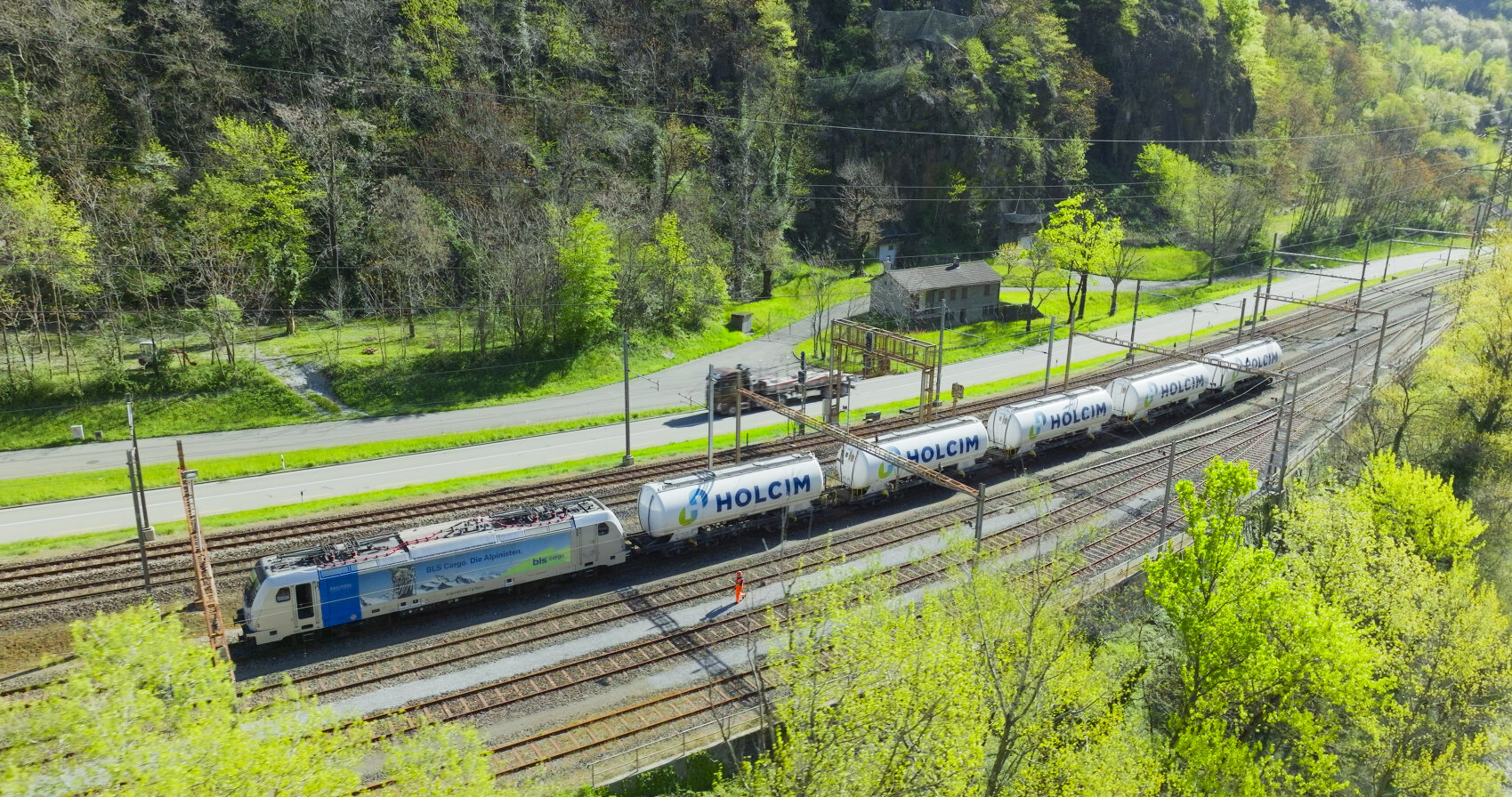 Ein Güterzug mit zylindrischen Kesselwagen der Marke Holcim fährt an einem sonnigen Tag entlang der Bahngleise neben einer Straße, umgeben von Bäumen und Grünflächen.