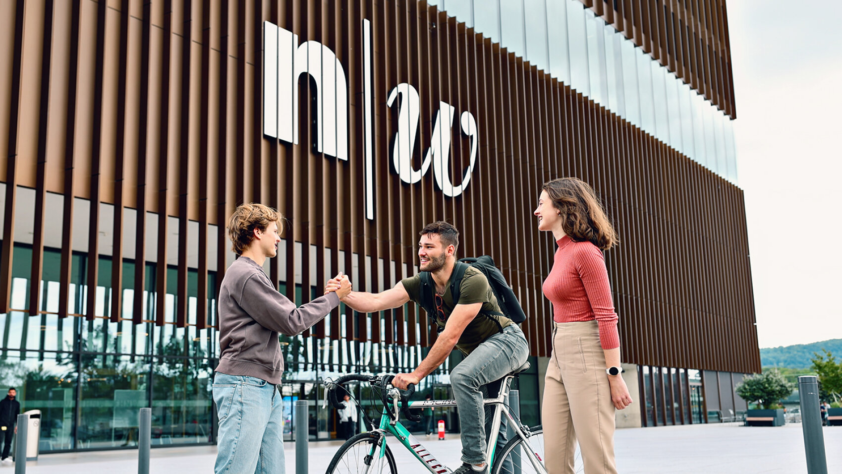 Drei junge Erwachsene begrüßen sich fröhlich vor einem modernen Gebäude mit großen n w-Buchstaben an der Fassade; einer hält ein Fahrrad in der Hand, und die anderen lächeln beim Händeschütteln.