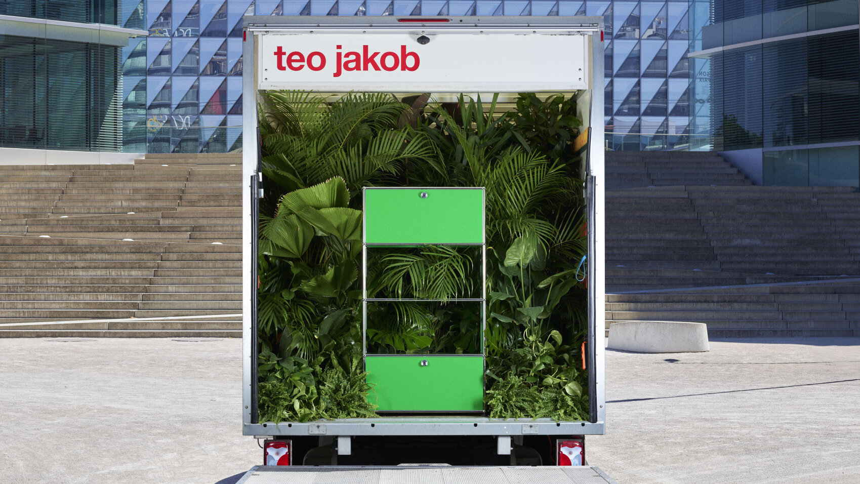 Un camion portant l'enseigne teo jakob est garé à l'extérieur, son arrière ouvert est rempli de plantes vertes luxuriantes et d'une armoire vert vif, le tout entouré d'un bâtiment moderne en verre et de larges marches en pierre.