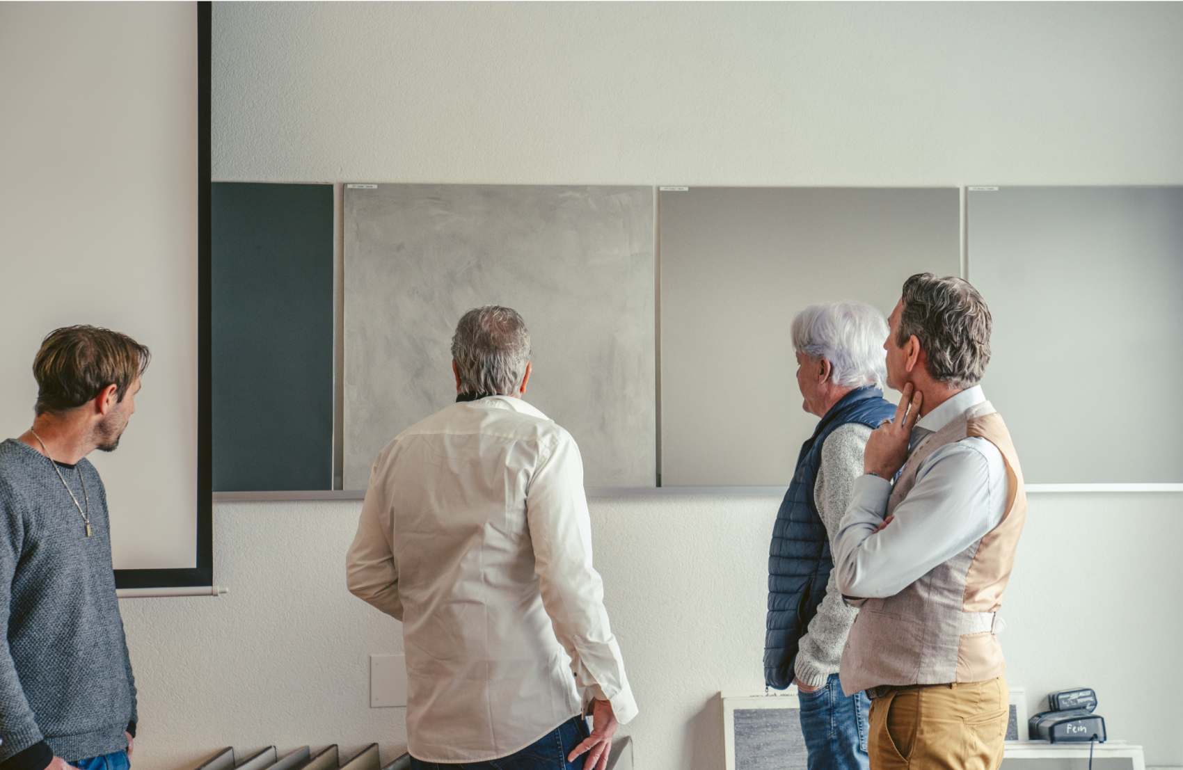 Quatre hommes se tiennent devant un tableau blanc vide dans une salle de classe ou un bureau et le regardent pensivement, le dos et les côtés tournés vers la caméra.