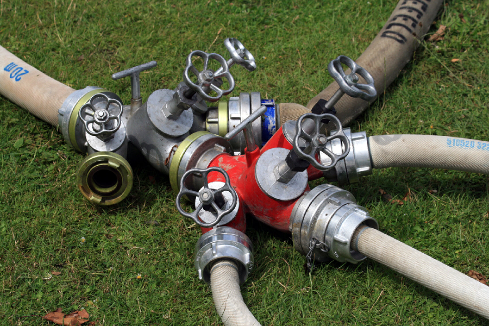Une prise d'eau avec plusieurs tuyaux, posée sur de l'herbe verte. Les vannes et les raccords métalliques sont visibles, deux tuyaux étant reliés par une section rouge et d'autres par des sections argentées.