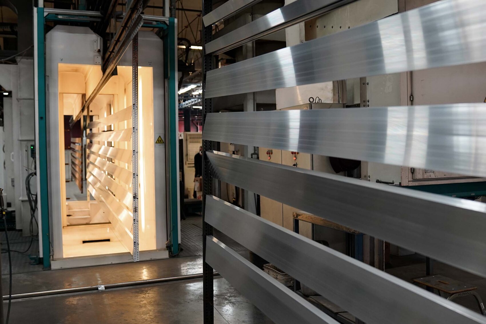 Des tôles métalliques brillantes sont positionnées devant une grande machine industrielle avec des lumières vives à l'intérieur, qui fait probablement partie d'un processus de fabrication ou d'affinage des métaux dans un environnement d'usine.