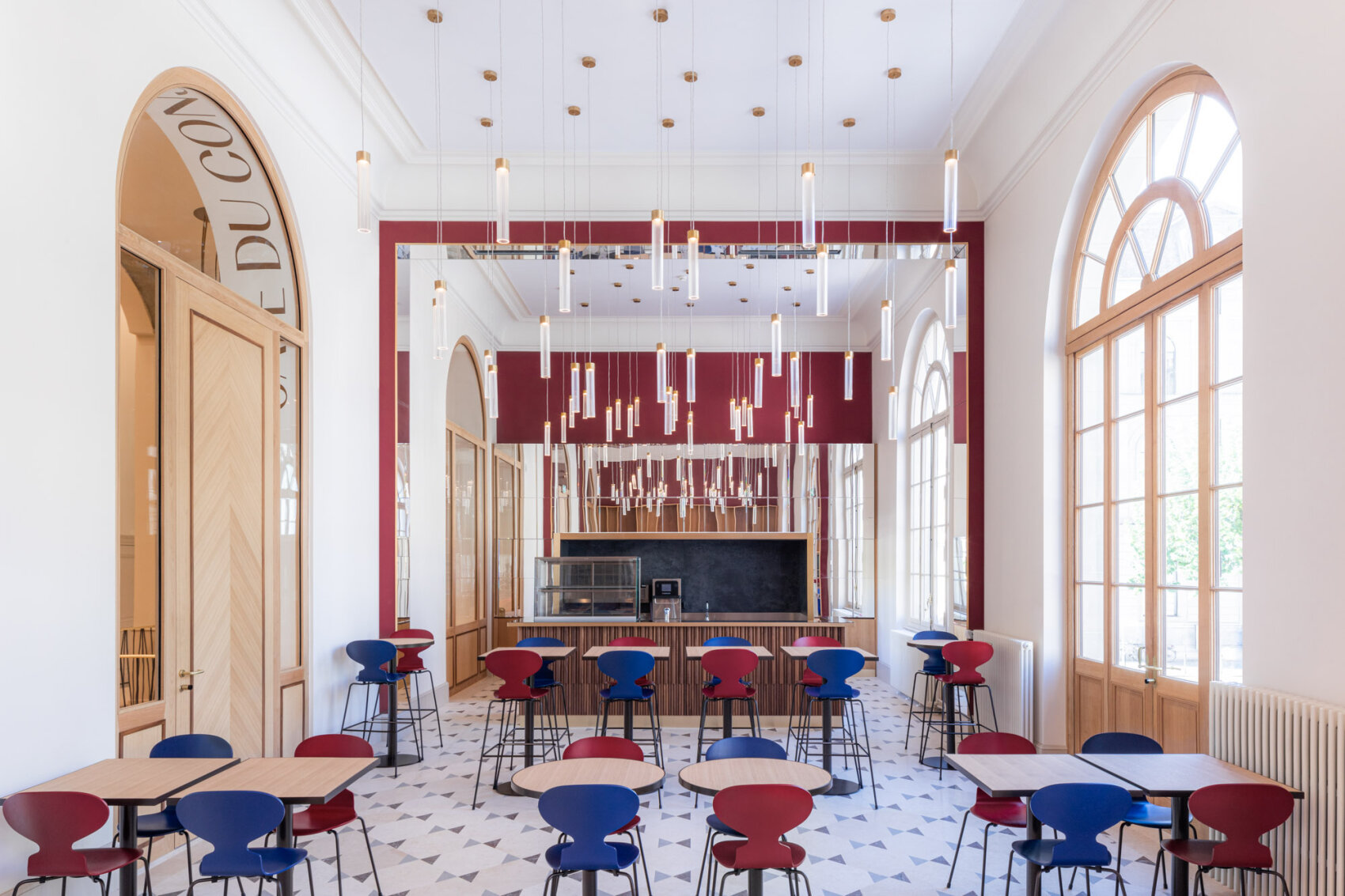 Café moderne avec de hauts plafonds, de grandes fenêtres cintrées et un sol carrelé géométrique. Des chaises bleues et rouges entourent des tables en bois clair. Des tubes lumineux suspendus et un panneau rouge en miroir accentuent l'espace minimaliste et aéré.