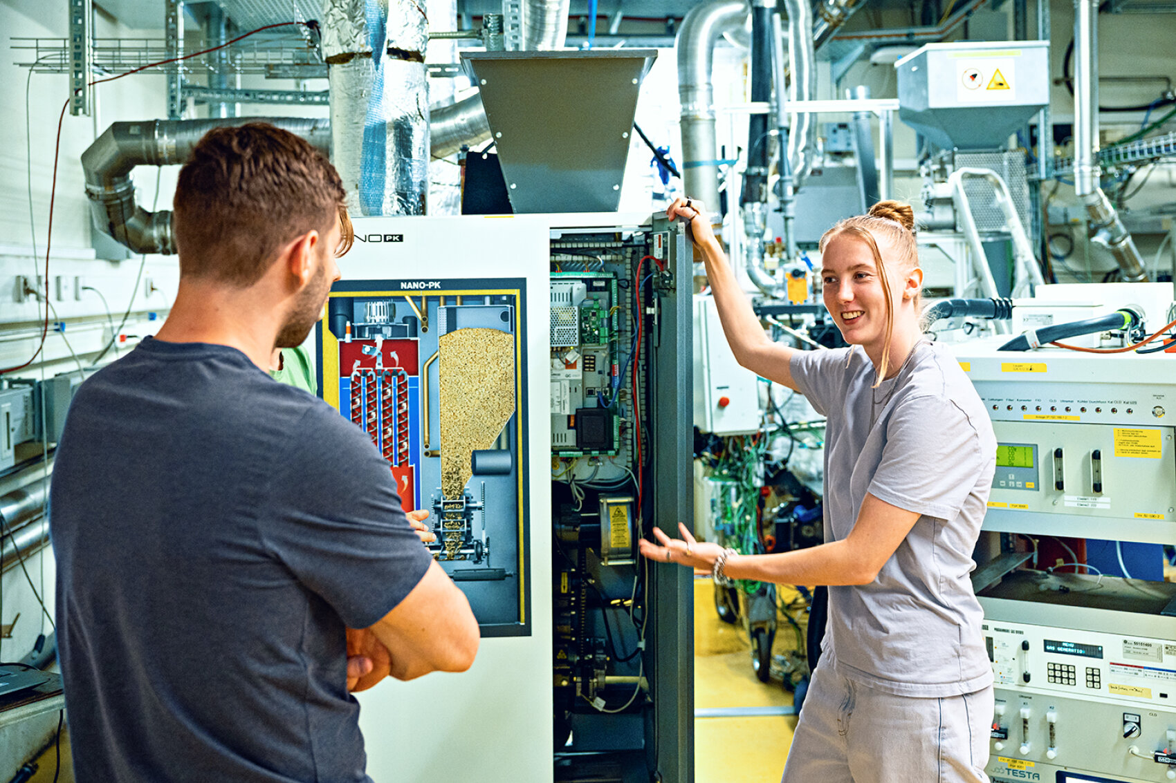 Eine Frau erklärt einem Mann in einem Industrielabor Maschinen und deutet auf ein Gerät mit freiliegenden Kabeln und einem Diagramm, umgeben von Rohren und technischen Geräten.