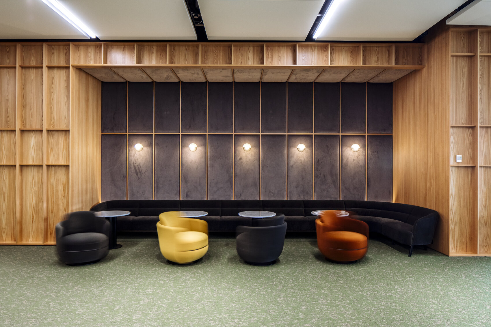 Salon moderne composé d'un canapé noir incurvé, de quatre chaises rondes noires, jaunes et orange, de petites tables rondes, de murs en bois, d'un mur d'accent foncé et de cinq lampes murales au-dessus des sièges.