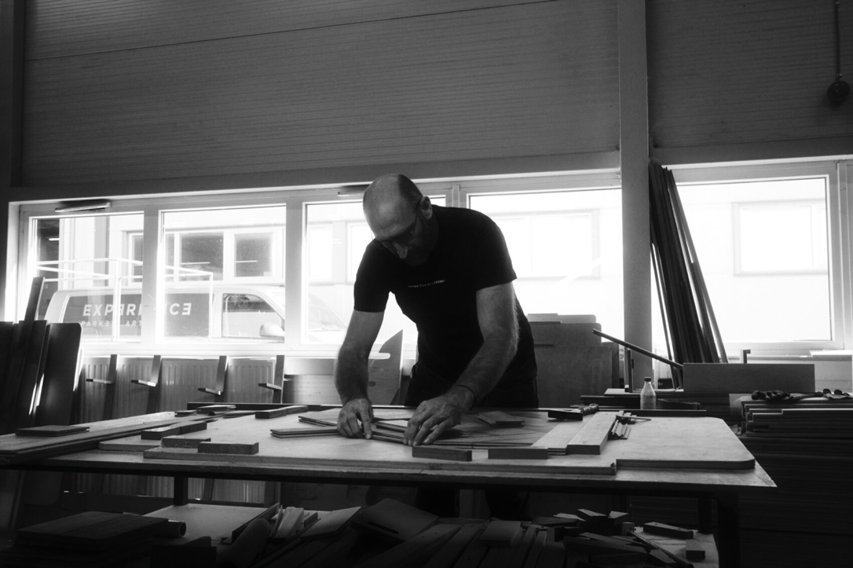 Un homme travaille avec des planches de bois sur une grande table dans un atelier, éclairé par la lumière naturelle qui passe par les fenêtres derrière lui.