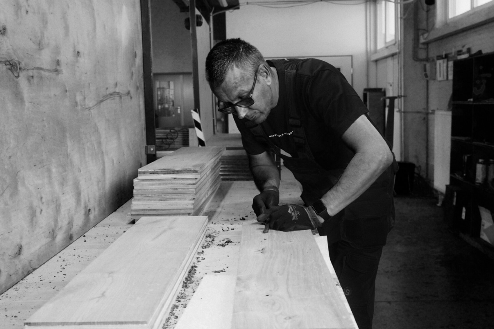 Un homme portant des lunettes et des gants travaille un morceau de bois sur un établi dans une menuiserie. Autour de lui, dans la pièce bien éclairée, on peut voir des piles de planches de bois et d'outils.