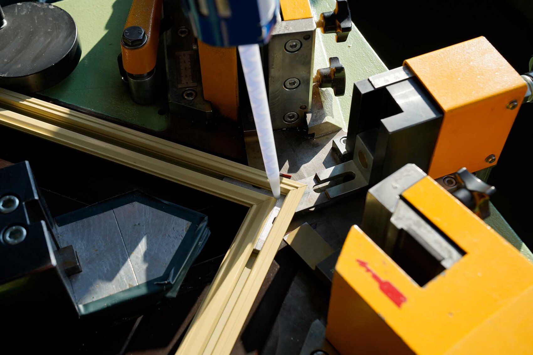 Gros plan sur une machine appliquant de la colle ou du mastic sur l'assemblage d'angle d'un cadre en bois, avec des pièces de machine jaunes et métalliques autour de la zone de travail.
