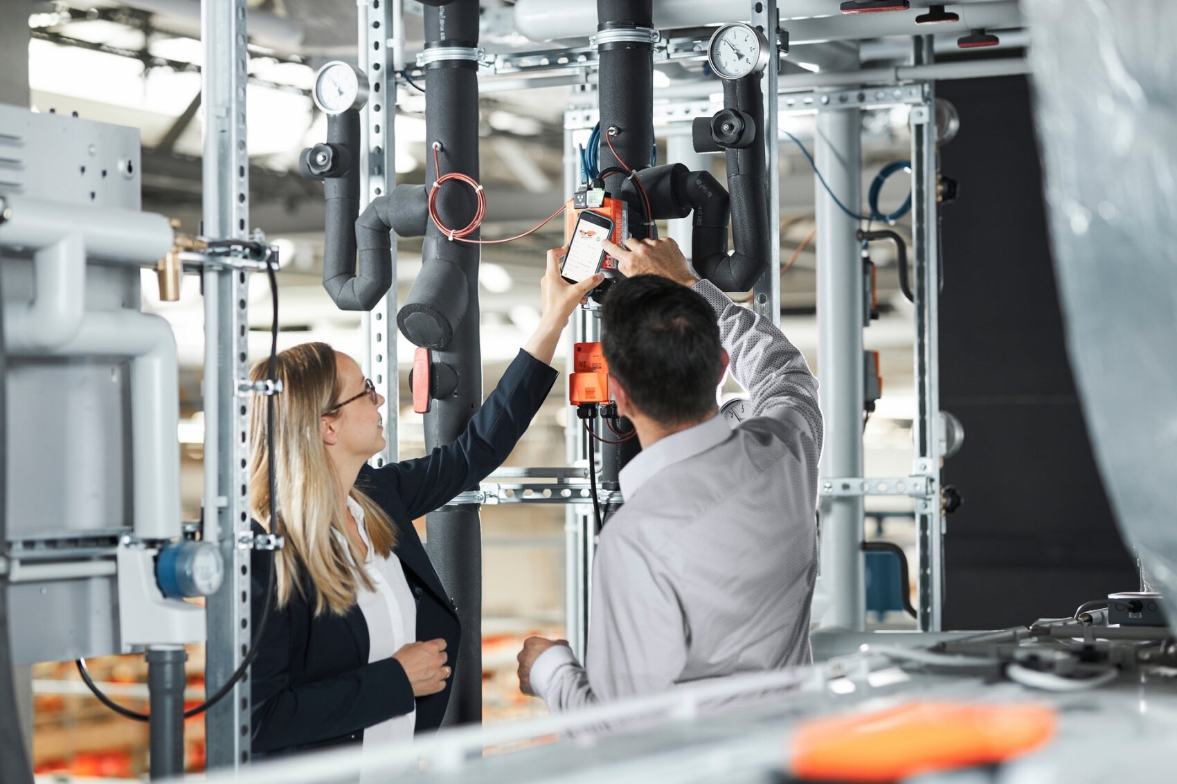 Zwei Personen, eine Frau und ein Mann, inspizieren und justieren Komponenten von Industrierohrleitungen in einer hell erleuchteten Anlage. Sie scheinen Messgeräte und Ventile im Rahmen einer Wartungs- oder Bewertungsaufgabe zu überprüfen.