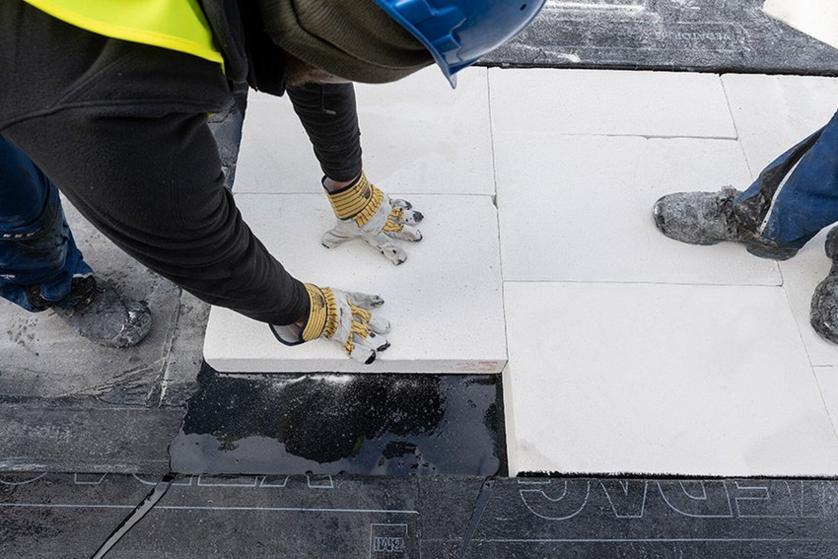 Un ouvrier du bâtiment portant des gants et un casque bleu pose des plaques de béton blanc sur le sol, tandis qu'une autre personne se tient à ses côtés, chaussée de bottes poussiéreuses.