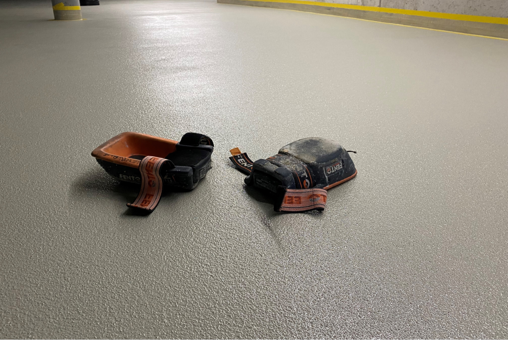 Deux genouillères usées noires et orange sont posées sur un sol structuré gris clair dans un intérieur avec un trottoir jaune en arrière-plan.